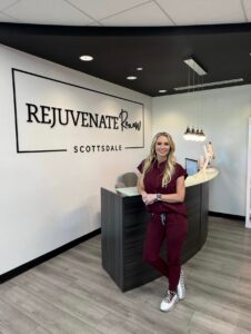 Cara Greer, CMLT, owner of Rejuvenate Renew, standing in front of the med spa's logo wall in Scottsdale, Arizona